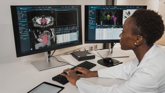 A radiologist sitting at a desk, looking at two computer screens showing scan software