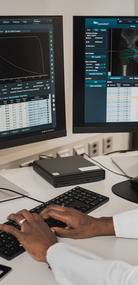 A radiologist sitting at a desk, looking at two computer screens showing scan software